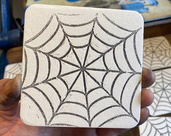 May include: A white square block with a black ink design of a spider web. The web has a central hub with radiating lines and concentric circles.