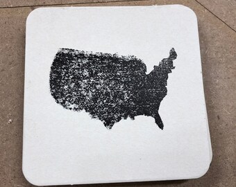 May include: A white square coaster with a black, textured outline of the United States. The coaster has rounded corners and is set against a neutral background. The design is simple and graphic.
