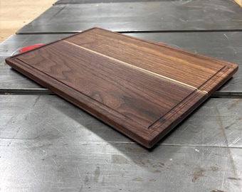 May include: A rectangular wooden cutting board with a dark brown finish and a lighter brown stripe down the center. The board has a carved edge and is likely used for food preparation. The board is approximately 12 inches long and 8 inches wide.