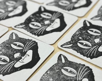 May include: A set of black and white cat coasters. Each coaster features a stylized illustration of a cat with a mischievous grin.