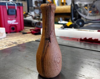 May include: A small, hand-turned wooden vase with a bulbous base and a narrow neck. The wood features a natural grain pattern with light brown and dark brown streaks, including a small, dark knot detail on the upper section of the neck.