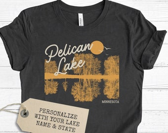 May include: A black t-shirt with a graphic of a lake scene with trees and a sun. The text "Pelican Lake" is written in a cursive font. The text "Minnesota" is written below the graphic. A tag is attached to the shirt that says "Personalize with your lake name & state".