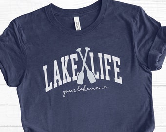 May include: A navy blue t-shirt with a white graphic of crossed paddles and the text "LAKE LIFE" in a white arch above the paddles. The text "your lake name" is written in white cursive below the paddles.