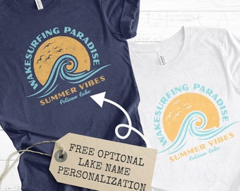 May include: Two t-shirts with a graphic design of a wave and the text "Wake Surfing Paradise Summer Vibes Pelican Lake". The text "Free Optional Lake Name Personalization" is on a brown tag.
