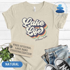 May include: A beige t-shirt with a retro-style graphic that says "Lake Life" with a rainbow design and the words "pelican lake" underneath. The shirt has a tag that says "Free Optional Lake Name Personalization" and the word "NATURAL" is printed on a tag below the shirt.