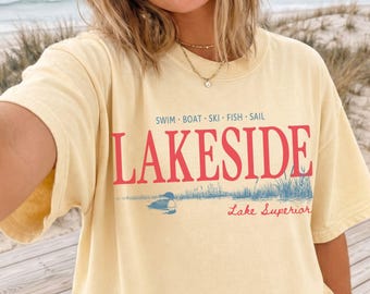 May include: A light yellow t-shirt with the word "LAKESIDE" in red block letters. Below the word is a blue and white graphic of a lake scene with the words "Lake Superior". Above the word "LAKESIDE" are the words "SWIM BOAT SKI FISH SAIL".