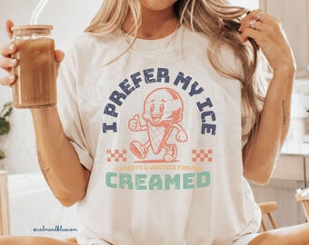May include: Cream-colored t-shirt with a retro ice cream cone graphic. The text on the shirt reads "I PREFER MY ICE CREAMED" in blue and green, with "LIBERTY & JUSTICE FOR ALL" in the middle.