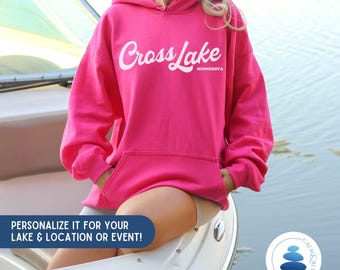 May include: A bright pink hooded sweatshirt with the text "Cross Lake Minnesota" in white script. The sweatshirt has a front pocket and a drawstring hood. The image also includes text that says "Personalize it for your lake & location or event!"