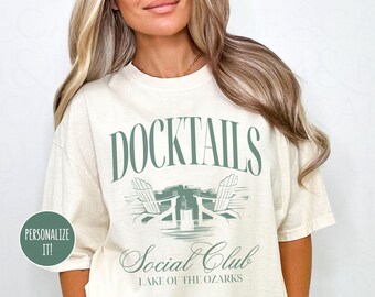 May include: A white t-shirt with a green graphic that says "Docktails Social Club Lake of the Ozarks". The graphic features two lounge chairs and a drink.
