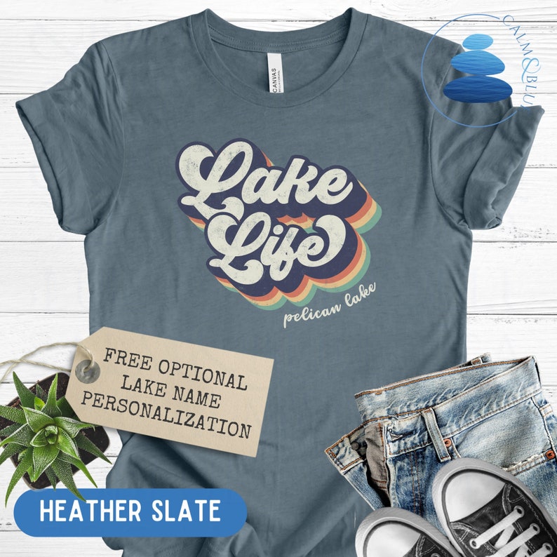 May include: Heather slate blue t-shirt with a retro-style graphic that says "Lake Life" with a rainbow design and the words "pelican lake" underneath. The t-shirt is shown on a white wooden background with a tag that says "Free Optional Lake Name Personalization".