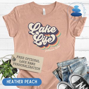 May include: A heather peach colored t-shirt with a retro-style graphic that says "Lake Life" with a rainbow design and the words "Pelican Lake" underneath. The shirt is displayed on a white wooden surface with a tag that says "Free Optional Lake Name Personalization".