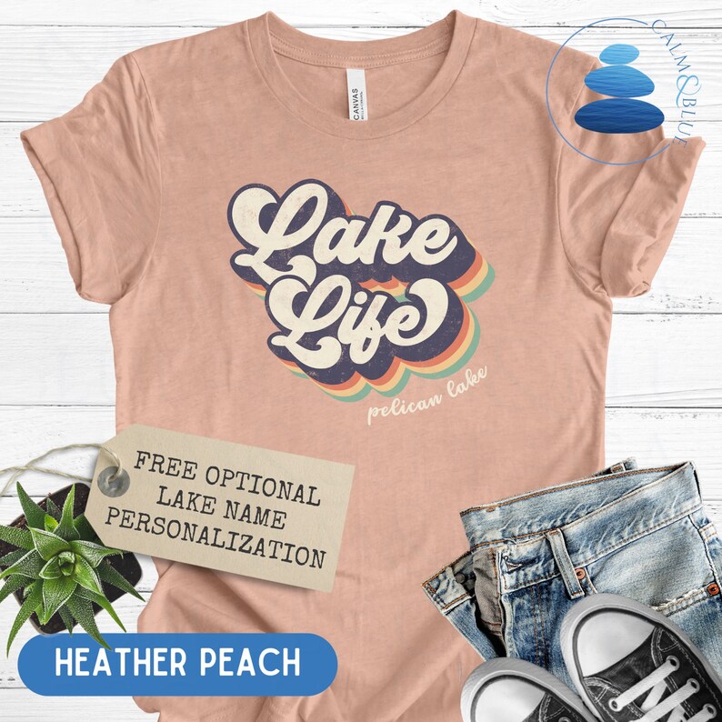 May include: A heather peach colored t-shirt with a retro-style graphic that says "Lake Life" with a rainbow design and the words "Pelican Lake" underneath. The shirt is displayed on a white wooden surface with a tag that says "Free Optional Lake Name Personalization".