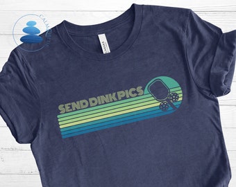May include: A navy blue t-shirt with a retro-style design featuring a pickleball paddle and the text "SEND DINK PICS" in yellow and green.