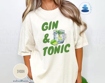 May include: Ivory-colored t-shirt with a green "Gin & Tonic" graphic. The design includes a cocktail illustration with lime wedges and ice, and a recipe. The shirt is a relaxed fit.