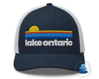 May include: A navy blue baseball cap with white mesh back panels. The front features an embroidered graphic of a yellow sun above horizontal stripes in orange, light blue, and dark blue, with the white embroidered text lake ontario below.