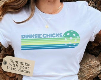 May include: White t-shirt with a retro design featuring the text "Dinksie Chicks" and a green and white pickleball. The design includes a blue and green striped pattern.