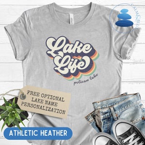 May include: A gray t-shirt with a retro-style graphic that says "Lake Life" with a rainbow design. The text "pelican lake" is printed below the graphic. The shirt is made of athletic heather fabric.