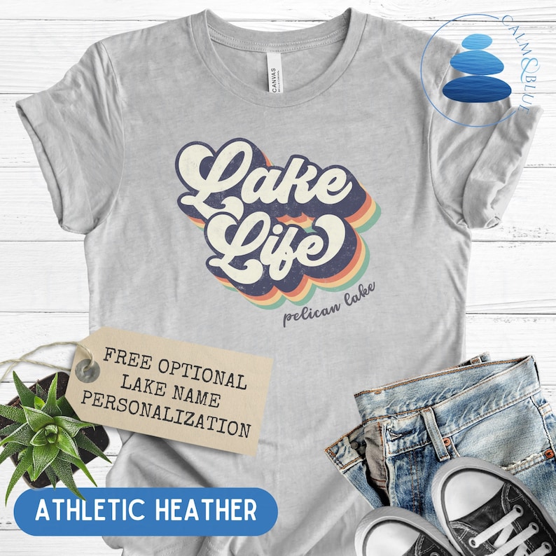 May include: A gray t-shirt with a retro-style graphic that says "Lake Life" with a rainbow design. The text "pelican lake" is printed below the graphic. The shirt is made of athletic heather fabric.