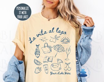 May include: Yellow t-shirt with a blue graphic design featuring the text "La vita al lago" and lake-themed illustrations. The design includes a fish, beach bag, book, umbrella, cocktail, and other summer elements. The text "Your Lake Here" is also included.