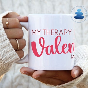 May include: White ceramic mug with a black handle. The mug has pink text that reads "MY THERAPY | Valen (A".