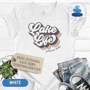 May include: White t-shirt with a retro-style graphic that says "Lake Life" with a rainbow design. The text "pelican lake" is written below the graphic. The shirt is displayed on a white wooden surface with a tag that says "Free Optional Lake Name Personalization".