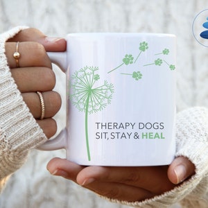 May include: White ceramic mug with a green dandelion design and the text "Therapy Dogs Sit, Stay & Heal".