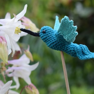 Op de afbeelding: Een handgemaakte, turquoise blauwe gebreide kolibrie op een houten stok, geplaatst in de buurt van witte bloemen met gele centra. De kolibrie heeft een lange zwarte snavel en gedetailleerde vleugels, tegen een vage groene achtergrond.