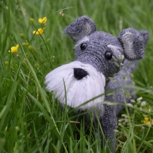Op de afbeelding: Een grijs gehaakt miniatuur schnauzer hondenspeelgoed met wit bont rond zijn gezicht en zwarte ogen. Het speelgoed zit in een veld met groen gras en gele bloemen.