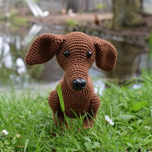 Op de afbeelding: Een bruin gehaakt speelgoed teckel dat in een veld met groen gras en witte bloemen zit.