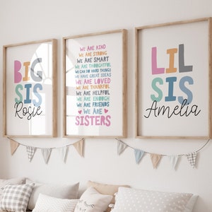 Peut inclure: Deux impressions encadrées avec du texte coloré. La première impression dit "BIG SIS Rosie" en couleurs arc-en-ciel. La deuxième impression dit "WE ARE KIND, WE ARE STRONG, WE ARE SMART, WE ARE THOUGHTFUL, WE CAN DO HARD THINGS, WE HAVE GREAT IDEAS, WE ARE LOVED, WE ARE THANKFUL, WE ARE HELPFUL, WE ARE ENOUGH, WE ARE FRIENDS, WE ARE SISTERS" en couleurs arc-en-ciel. La troisième impression dit "LIL SIS Amelia" en couleurs arc-en-ciel.