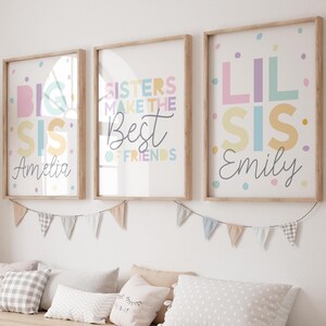 May include: Three framed art prints with pastel text and confetti. The prints read "Big Sis Amelia", "Sisters Make The Best Of Friends", and "Lil Sis Emily". A garland of small flags hangs below the prints, and pillows are on a bench below.