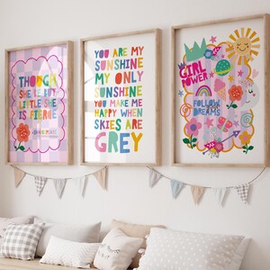 May include: Three framed art prints with motivational quotes and colourful illustrations. The prints feature phrases like "You are my sunshine" and "Girl Power." The frames are light wood, displayed above a bench with pillows.