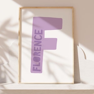 Op de afbeelding: Een ingelijst print met een lichtpaarse letter F en het woord "FLORENCE" dat verticaal in de letter is geschreven. De print is op een witte achtergrond en is ingelijst in een licht houten lijst.