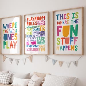 Puede incluir: Tres impresiones artísticas enmarcadas con texto colorido: "Where the Wild Ones Play", "Playroom Rules" y "This is Where the Fun Stuff Happens". Las impresiones se muestran encima de una unidad de almacenamiento de madera con contenedores blancos y almohadas decorativas.