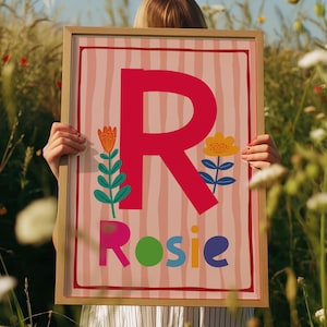 May include: A framed art print featuring a large red letter "R" with the name "Rosie" below. The background is pink with vertical stripes, and the artwork includes floral illustrations. The frame is a light wood color.