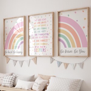May include: Three framed wall art prints featuring pastel rainbows and inspirational text including "Be kind Be happy," "We are sisters," and "Be brave Be You." The prints are displayed above a wooden storage unit with white and beige bins.