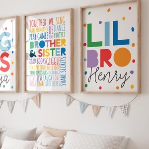 May include: Three colourful framed prints with the words "Big Sis Maya", "Lil Bro Henry", and a list of things siblings do together. The prints are on a white wall with a wooden shelf below.