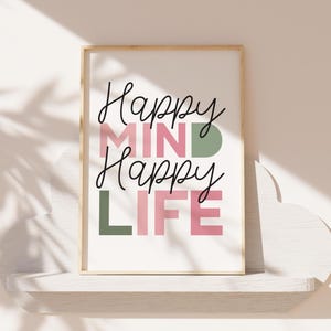 May include: A framed print with the words "Happy MIND Happy LIFE" in black script and block letters. The word "MIND" is in pink, and the "D" is green. The word "LIFE" is in pink, and the "L" is green. The frame is a light wood color.