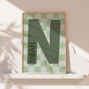 Op de afbeelding: Een ingelijste print met een grote, olijfgroene letter "N" tegen een lichtgroene en witte geblokte achtergrond. De naam "NOAH" is verticaal aan de linkerkant van de letter afgedrukt. De lijst heeft een lichte houtkleur.