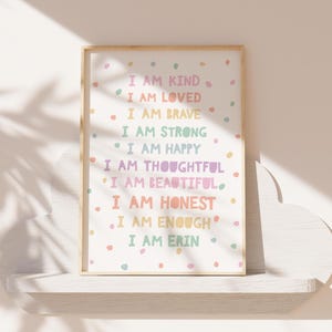 May include: A framed print with a white background and colorful polka dots. The print features a list of positive affirmations in a rainbow color scheme, including "I am kind", "I am loved", "I am brave", "I am strong", "I am happy", "I am thoughtful", "I am beautiful", "I am honest", "I am enough", and "I am Erin".