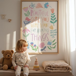 May include: A framed print with the words "BEAUTIFUL GIRL YOU WERE MADE FOR AMAZING THINGS" in colorful lettering and floral illustrations. A young child in a daisy-print outfit sits on a bench with a teddy bear, a wooden stacking toy, and a folded blanket.