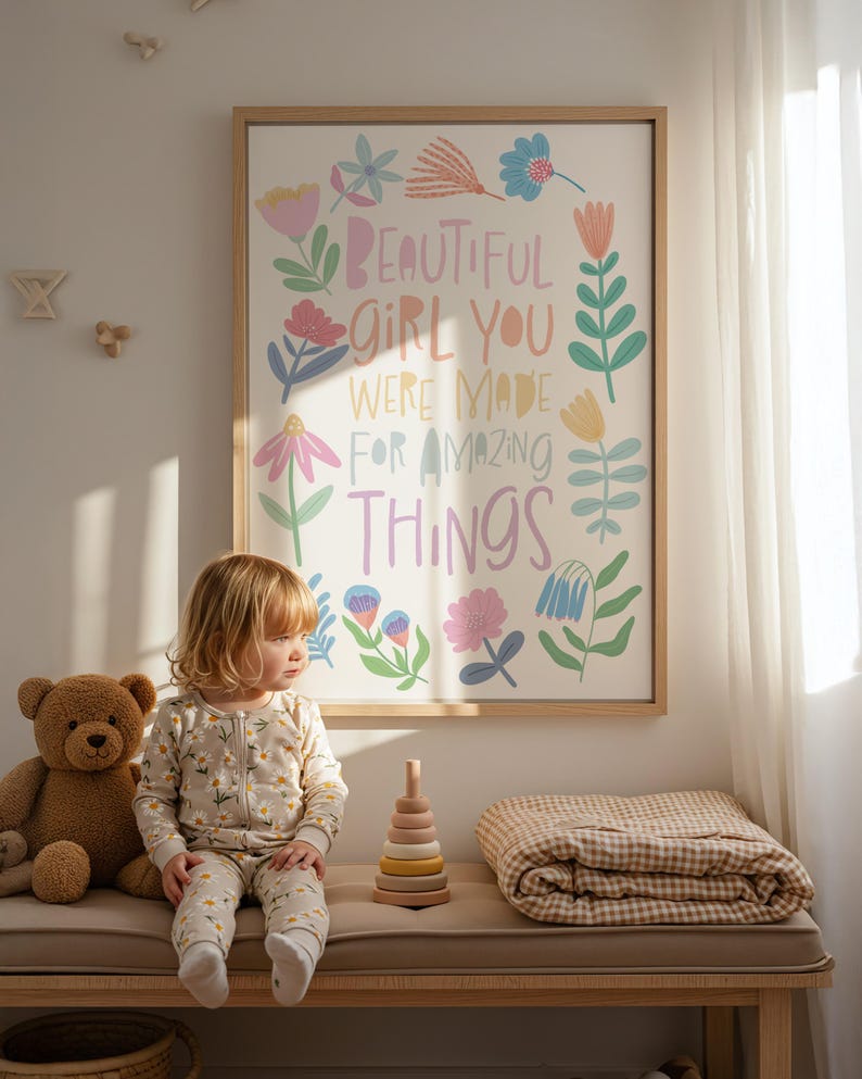 May include: A framed print with the words "BEAUTIFUL GIRL YOU WERE MADE FOR AMAZING THINGS" in colorful lettering and floral illustrations. A young child in a daisy-print outfit sits on a bench with a teddy bear, a wooden stacking toy, and a folded blanket.