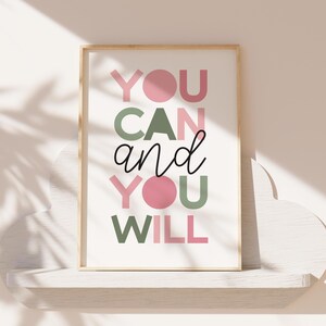 May include: A framed print with the words "YOU CAN and YOU WILL" in pink, green, and black lettering. The artwork has a light wood frame and is displayed on a white shelf against a neutral background.