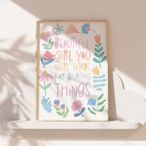 May include: A framed art print with the words "BEAUTIFUL GIRL YOU WERE MADE FOR AMAZING THINGS" surrounded by colorful floral illustrations. The artwork is displayed on a white shelf against a neutral wall.