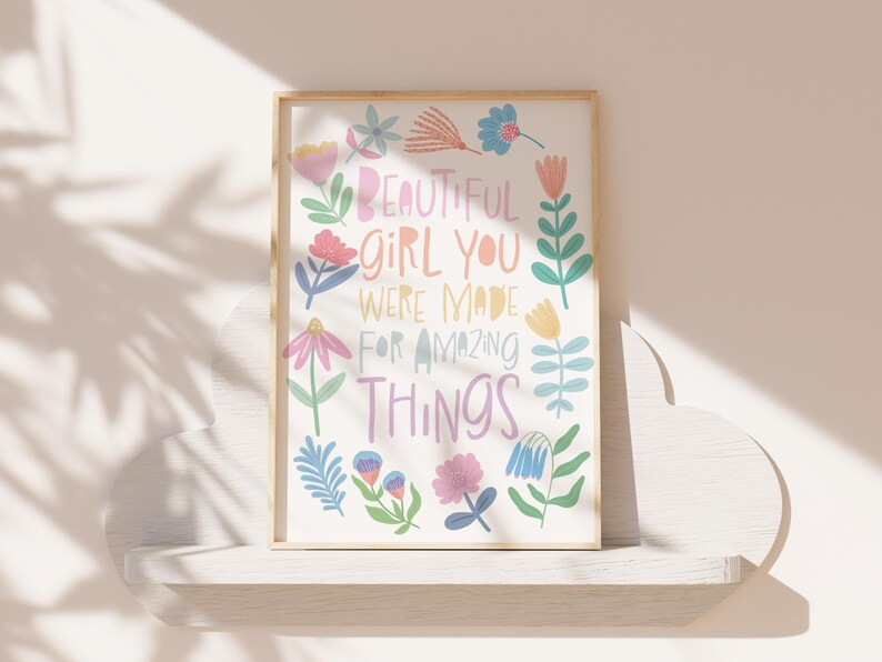 May include: A framed art print with the words "BEAUTIFUL GIRL YOU WERE MADE FOR AMAZING THINGS" surrounded by colorful floral illustrations. The artwork is displayed on a white shelf against a neutral wall.
