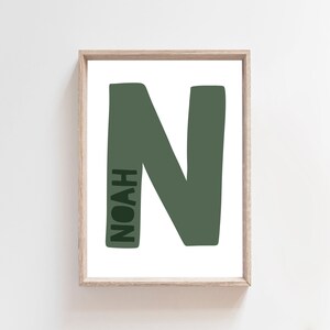 Op de afbeelding: Een ingelijst print met een grote, groene letter "N" en de naam "NOAH" die verticaal in de letter is geschreven.
