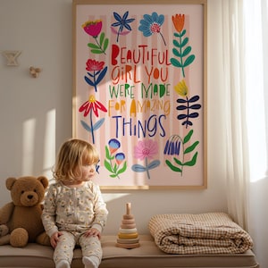 May include: A framed art print with the text "BEAUTIFUL GIRL YOU WERE MADE FOR AMAZING THINGS" in colorful letters, surrounded by floral illustrations. A child in a floral onesie sits on a bench next to a teddy bear and a wooden stacking toy.