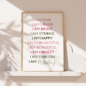 May include: A framed print with the words "I AM KIND, I AM LOVED, I AM BRAVE, I AM STRONG, I AM HAPPY, I AM THOUGHTFUL, I AM BEAUTIFUL, I AM HONEST, I AM ENOUGH, I AM Marley" in pink and green lettering. The print is in a light wood frame.