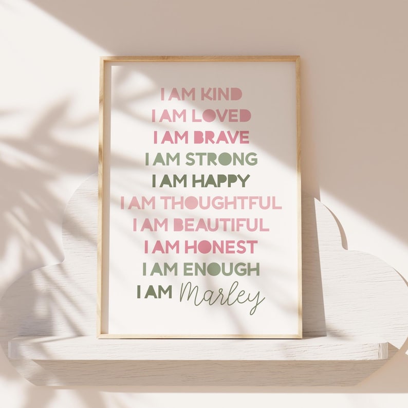May include: A framed print with the words "I AM KIND, I AM LOVED, I AM BRAVE, I AM STRONG, I AM HAPPY, I AM THOUGHTFUL, I AM BEAUTIFUL, I AM HONEST, I AM ENOUGH, I AM Marley" in pink and green lettering. The print is in a light wood frame.