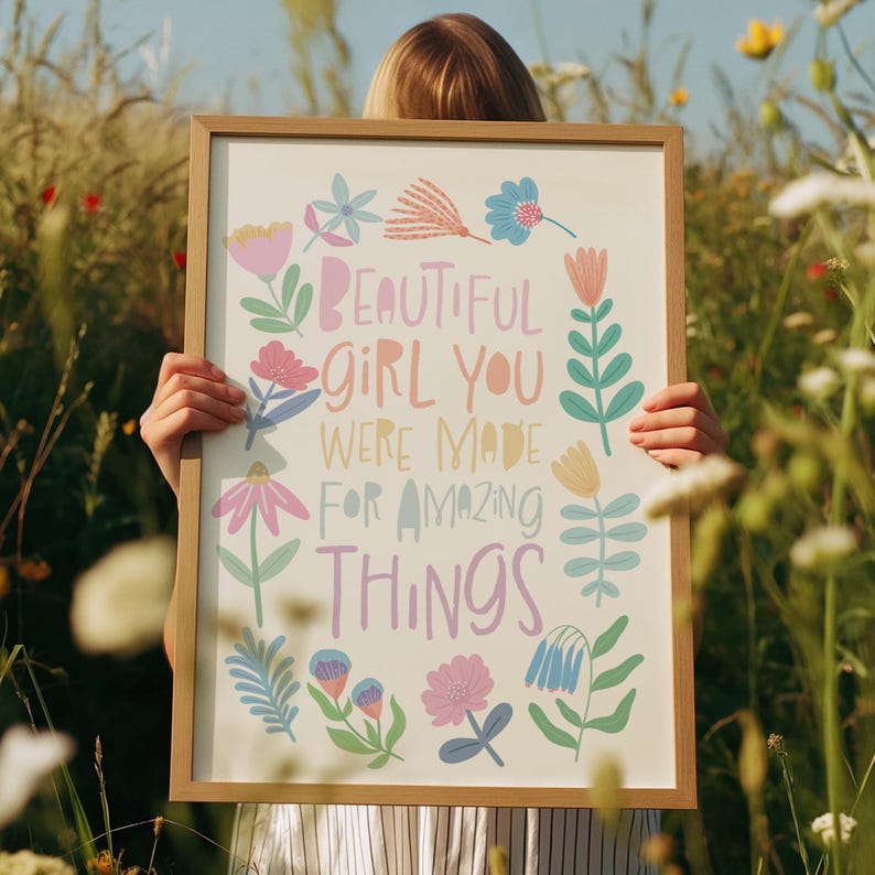 May include: A framed art print with the text "BEAUTIFUL GIRL YOU WERE MADE FOR AMAZING THINGS" in pastel colors. The print is surrounded by colorful floral illustrations and held by a person in a field of wildflowers.